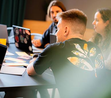 Drei Personen im Gespräch an einem Tisch mit Laptops und Papieren, im Fokus eine Person, die ein schwarzes Shirt mit einem bunten Grafikmotiv trägt