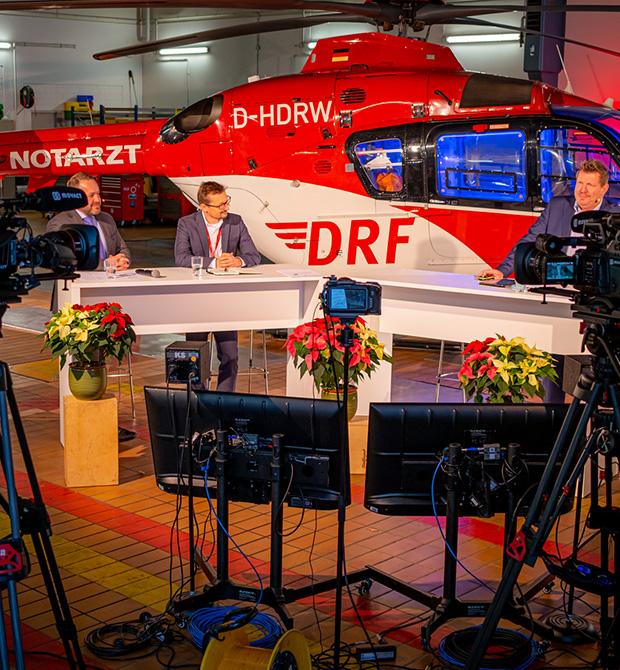 A panel discussion in front of a red and white DRF helicopter, with cameras and monitors set up for recording.