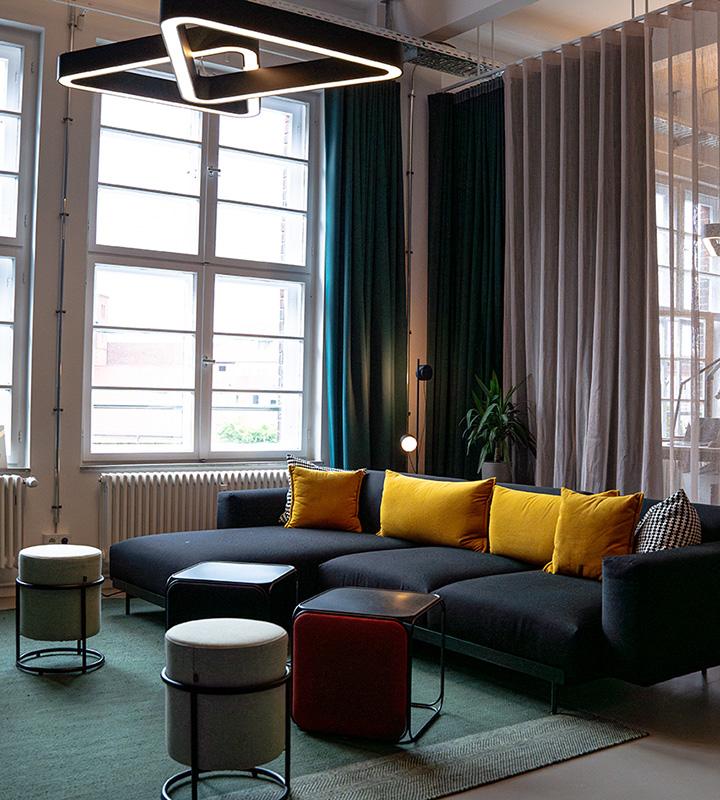 Movact.studio Eingangsbereich, gemütlicher Loungebereich mit einem großen schwarzen Sofa, bunten Kissen und einem Couchtisch vor großen Fenstern mit Vorhängen