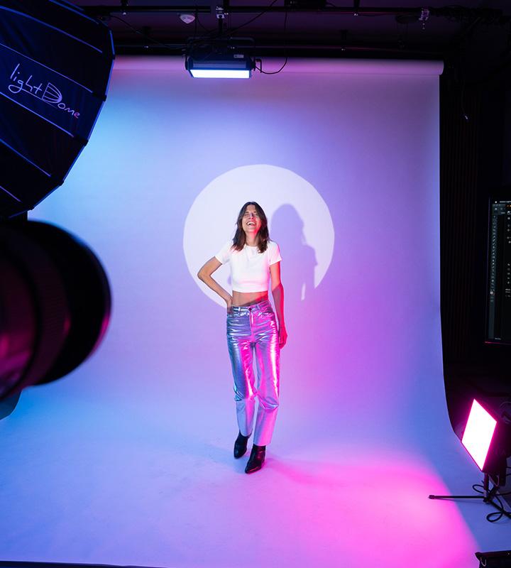 Fashion shoot with a woman in a white crop top and metallic pants, standing under blue and pink lights.