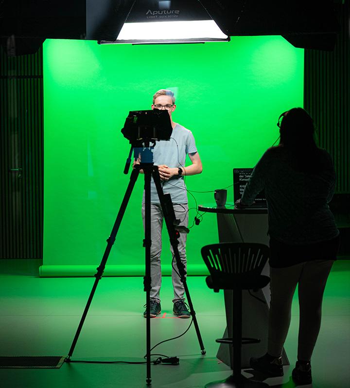 Person standing in front of a green screen with a camera and lighting setup, and another person operating equipment.