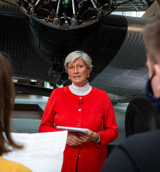 A woman speaking in front of an aircraft engine, holding documents, with two people in the foreground.