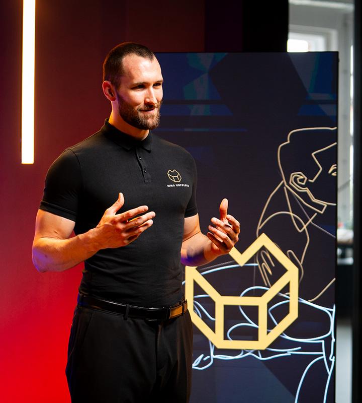 Man giving a presentation, wearing a black polo shirt with a logo, and gesturing with his hands.